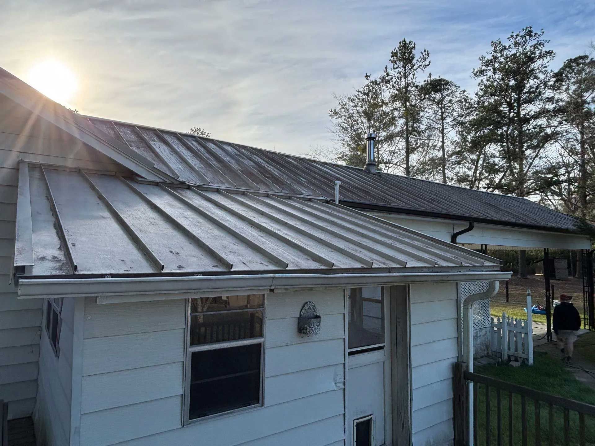 White metal roof on a house with a porch. Sun shines in the background. A person walks on a pathway.