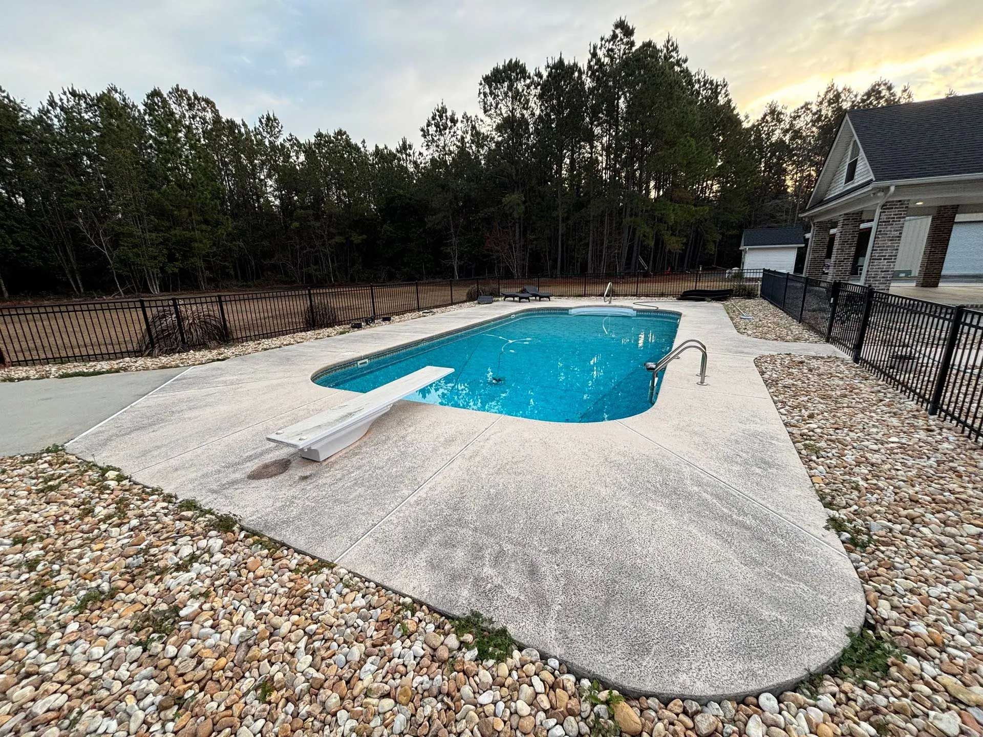 A swimming pool with a diving board in a backyard, surrounded by concrete and gravel.