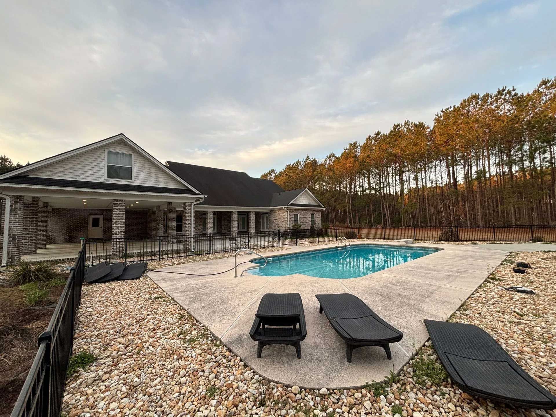 House with a pool and lounge chairs on a stone patio, surrounded by a black fence and trees.