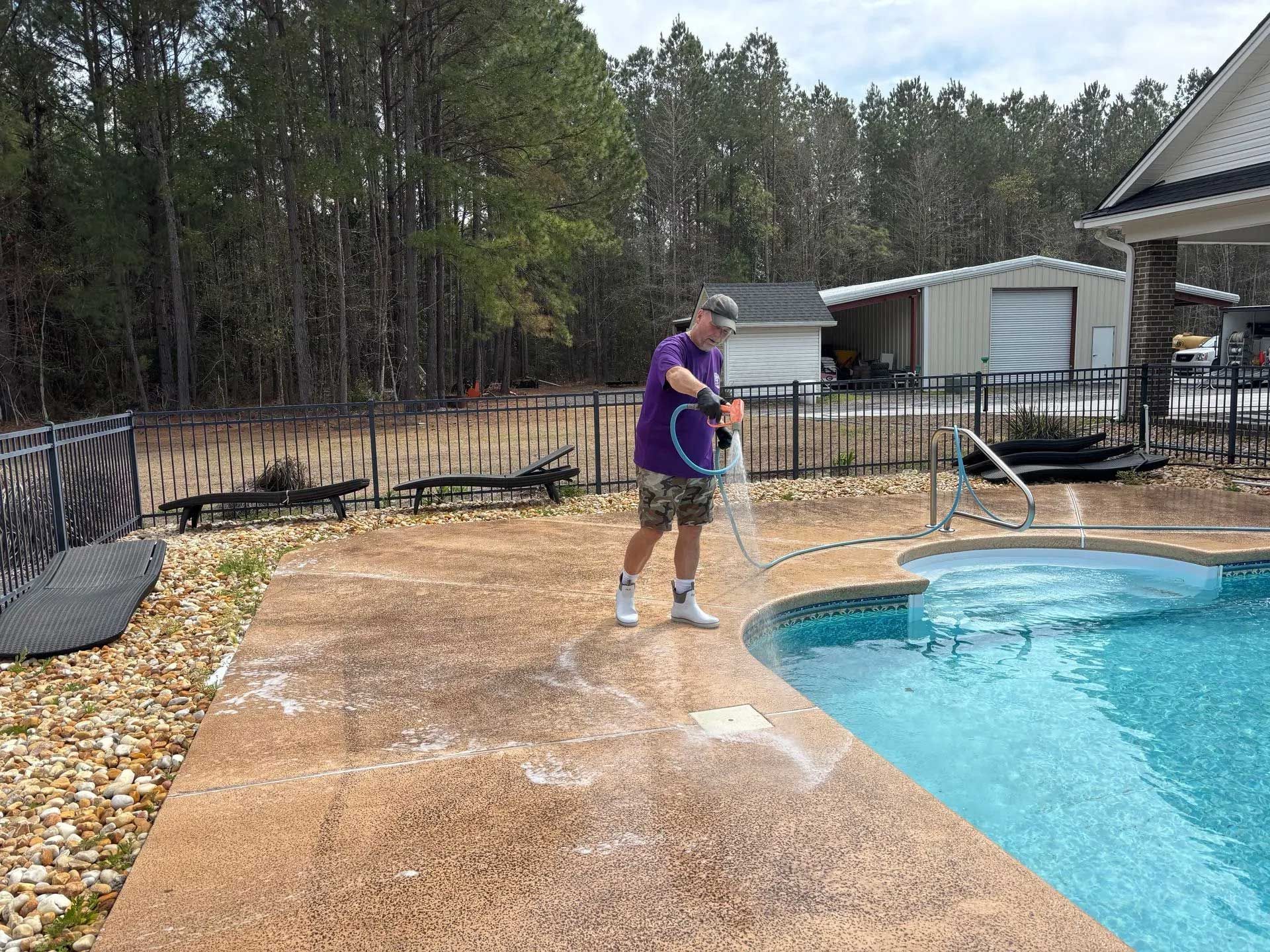 Person sprays a concrete patio near a pool with a hose, in a backyard setting.