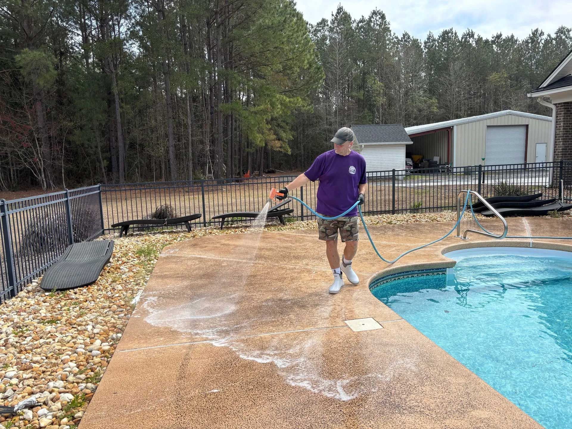 Man sprays water on a concrete pool deck near the pool. Trees and a small building are in the background.