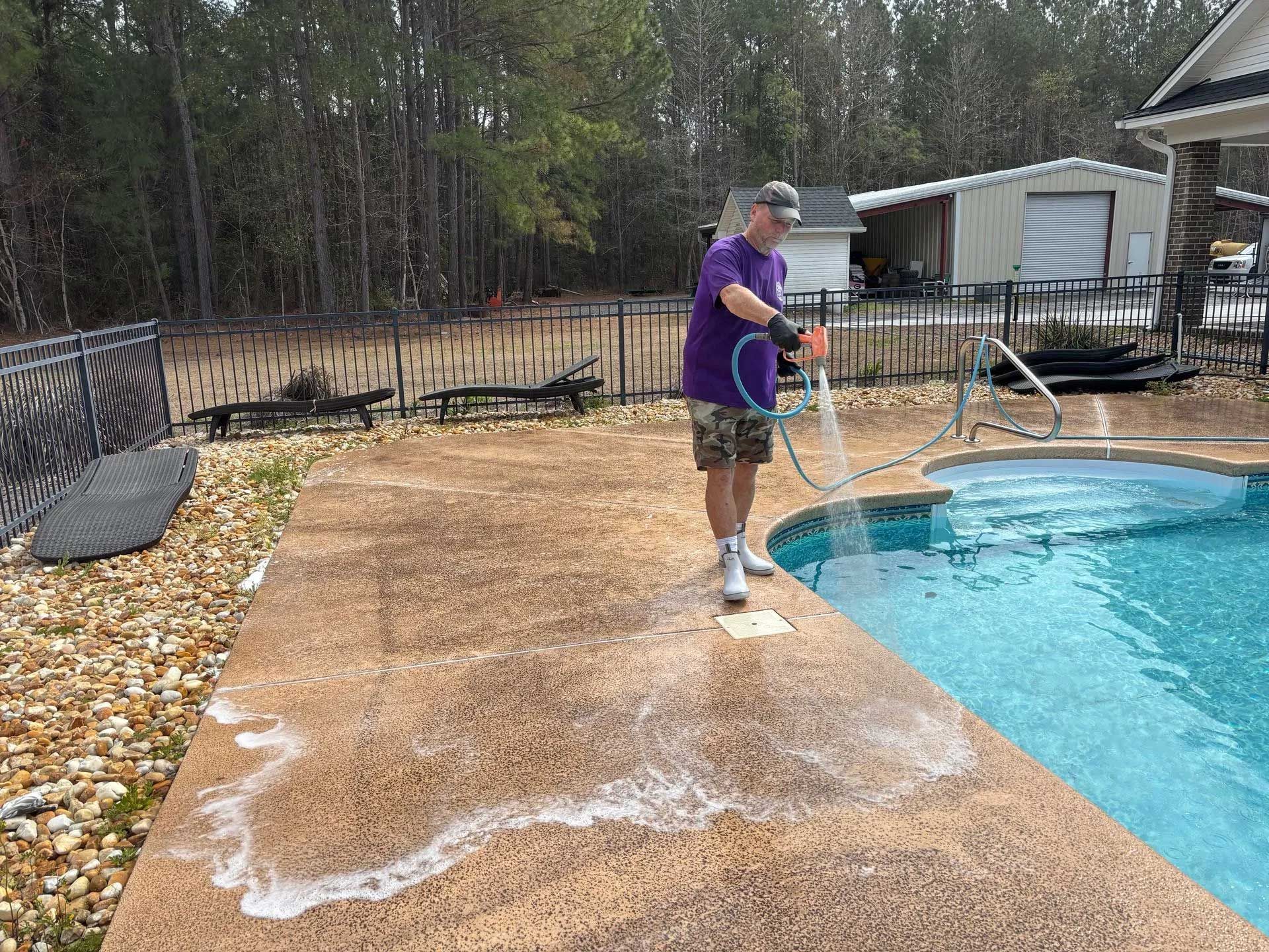 Man in purple shirt sprays concrete pool deck with water.