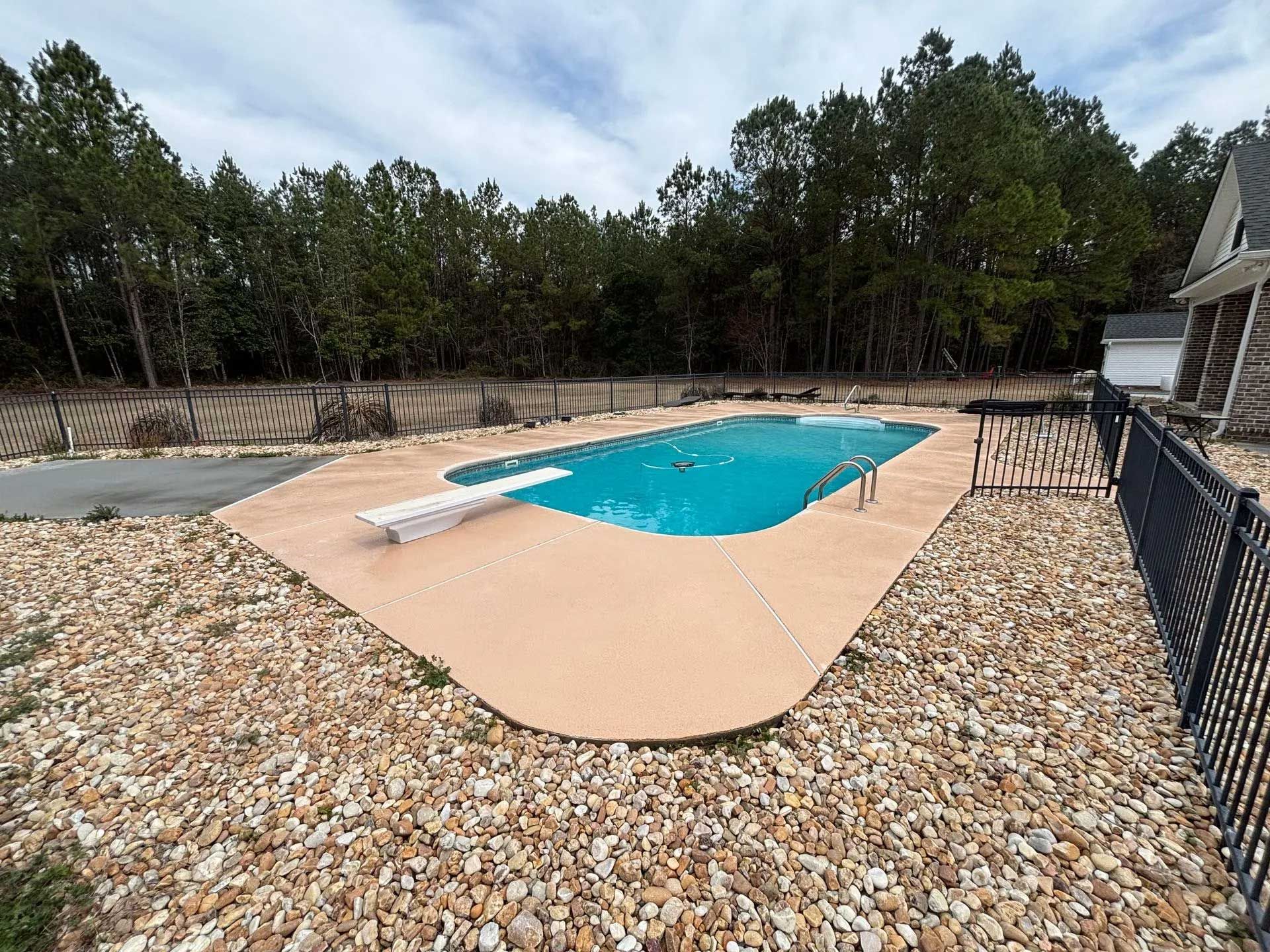 A rectangular swimming pool surrounded by tan concrete and rocks, with a diving board.