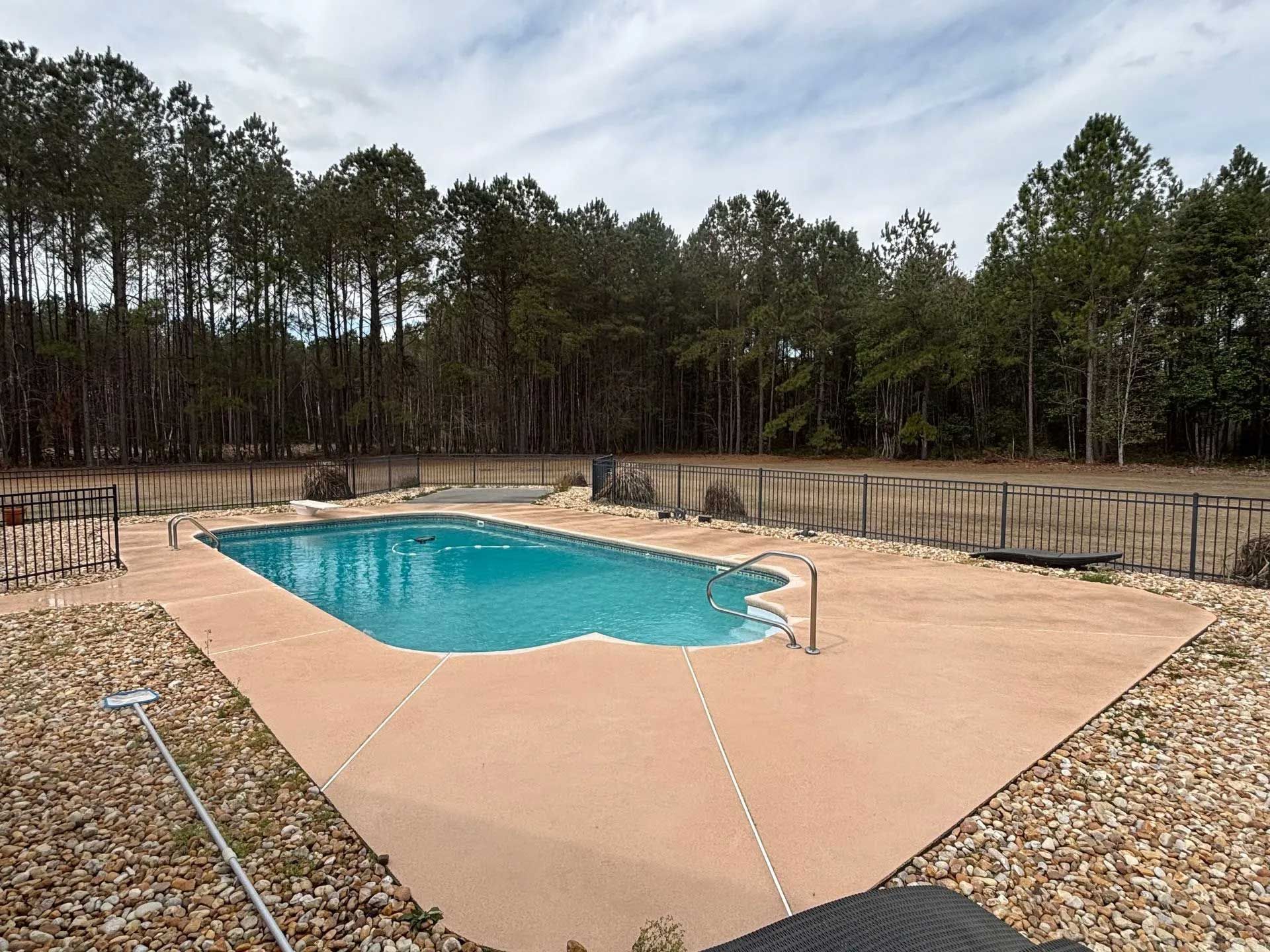 Pool with light blue water and tan concrete surround, backed by trees and a fence.