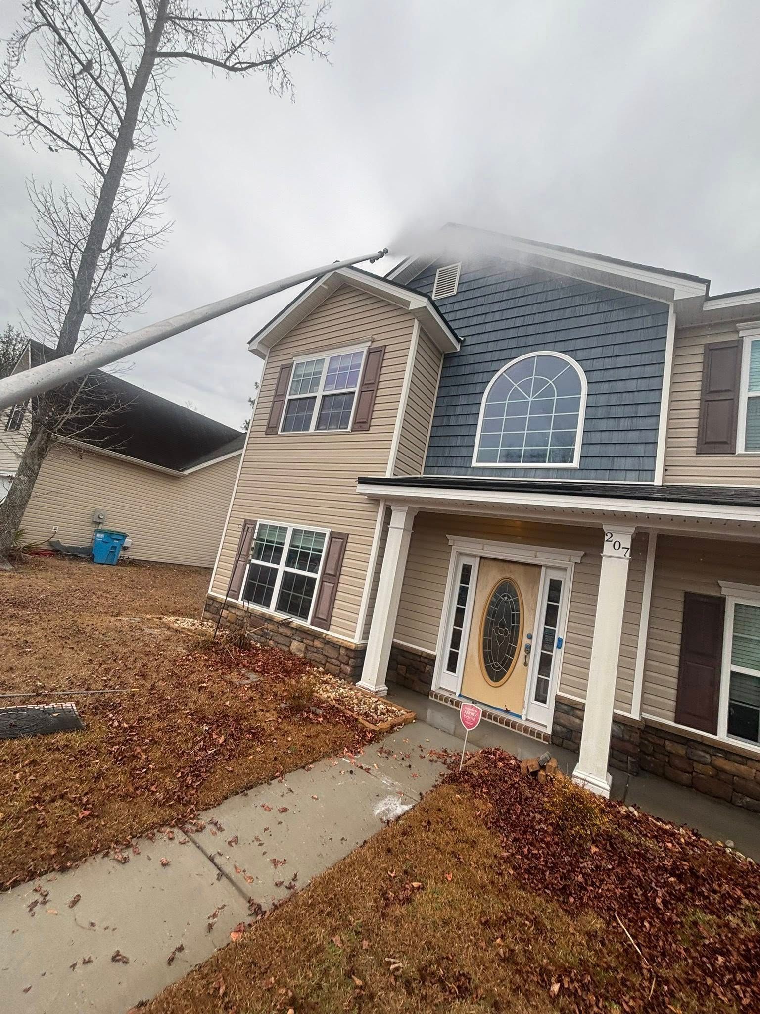 A worker using a long pole to spray-clean the dark roof shingles of a multi-story house with tan vinyl siding.