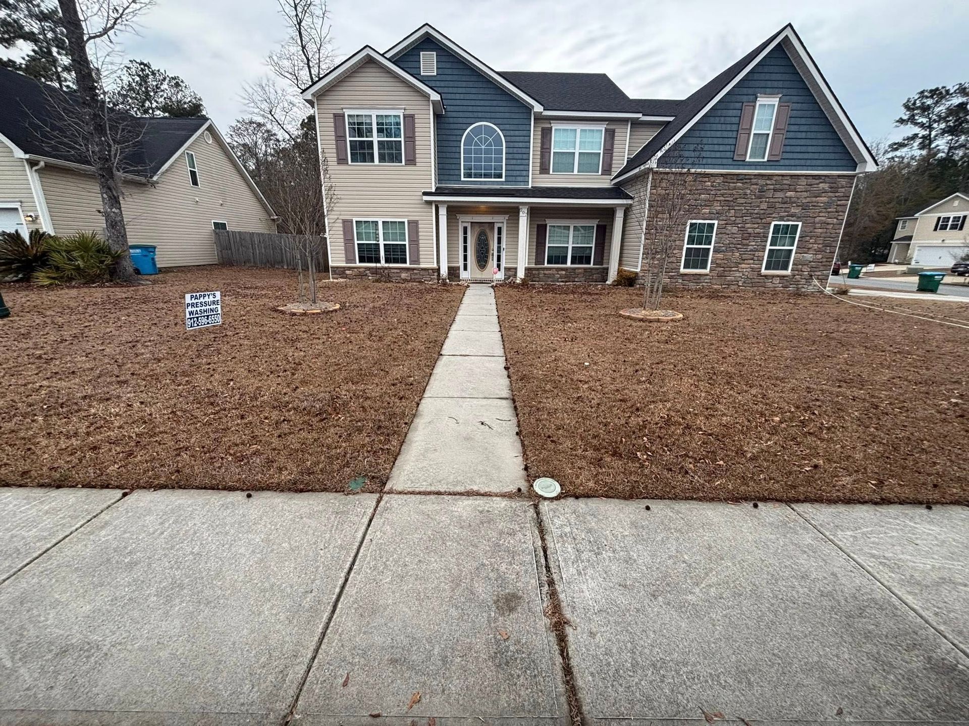A two-story suburban house with beige siding, dark blue accents, and a stone facade, featuring a lawn covered in brown leaves.