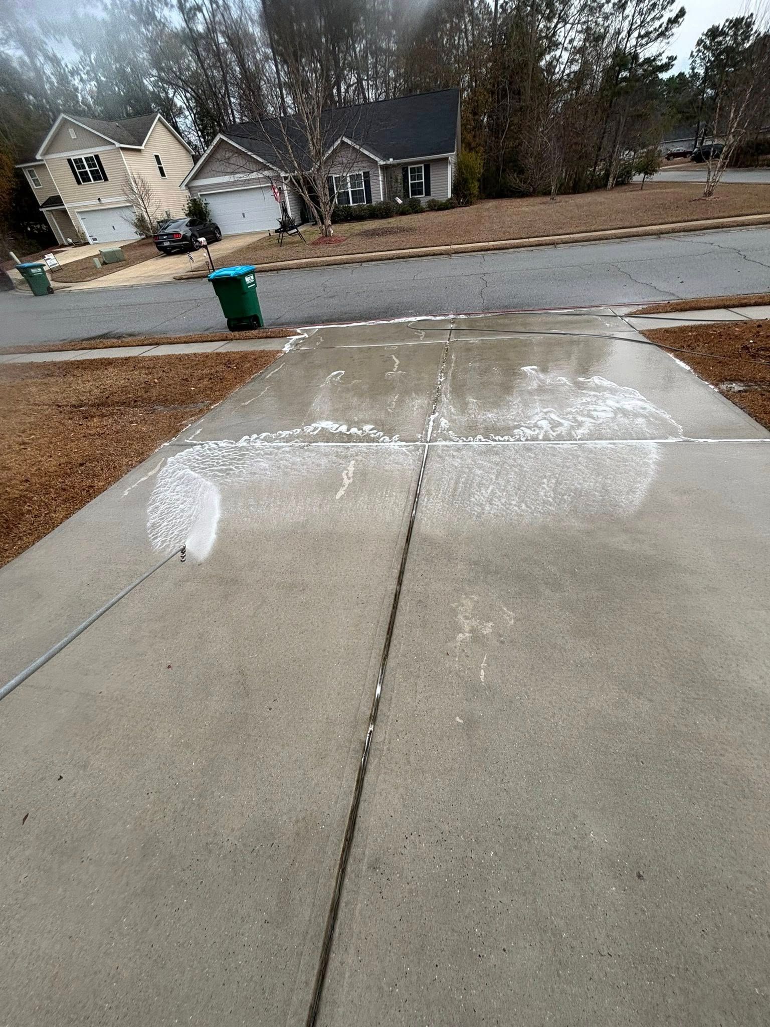 A concrete driveway with a large, white, foamy patch of cleaner drying on the surface, with houses in the background.