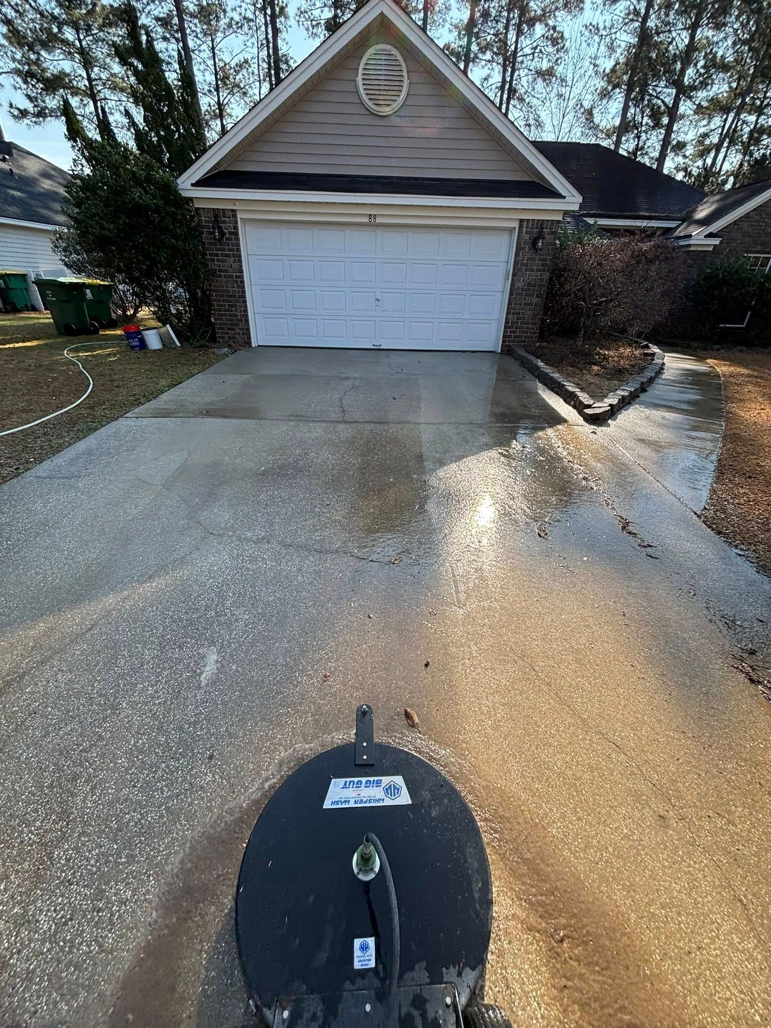 A driveway cleaning tool sits on a wet concrete driveway in front of a residential garage.