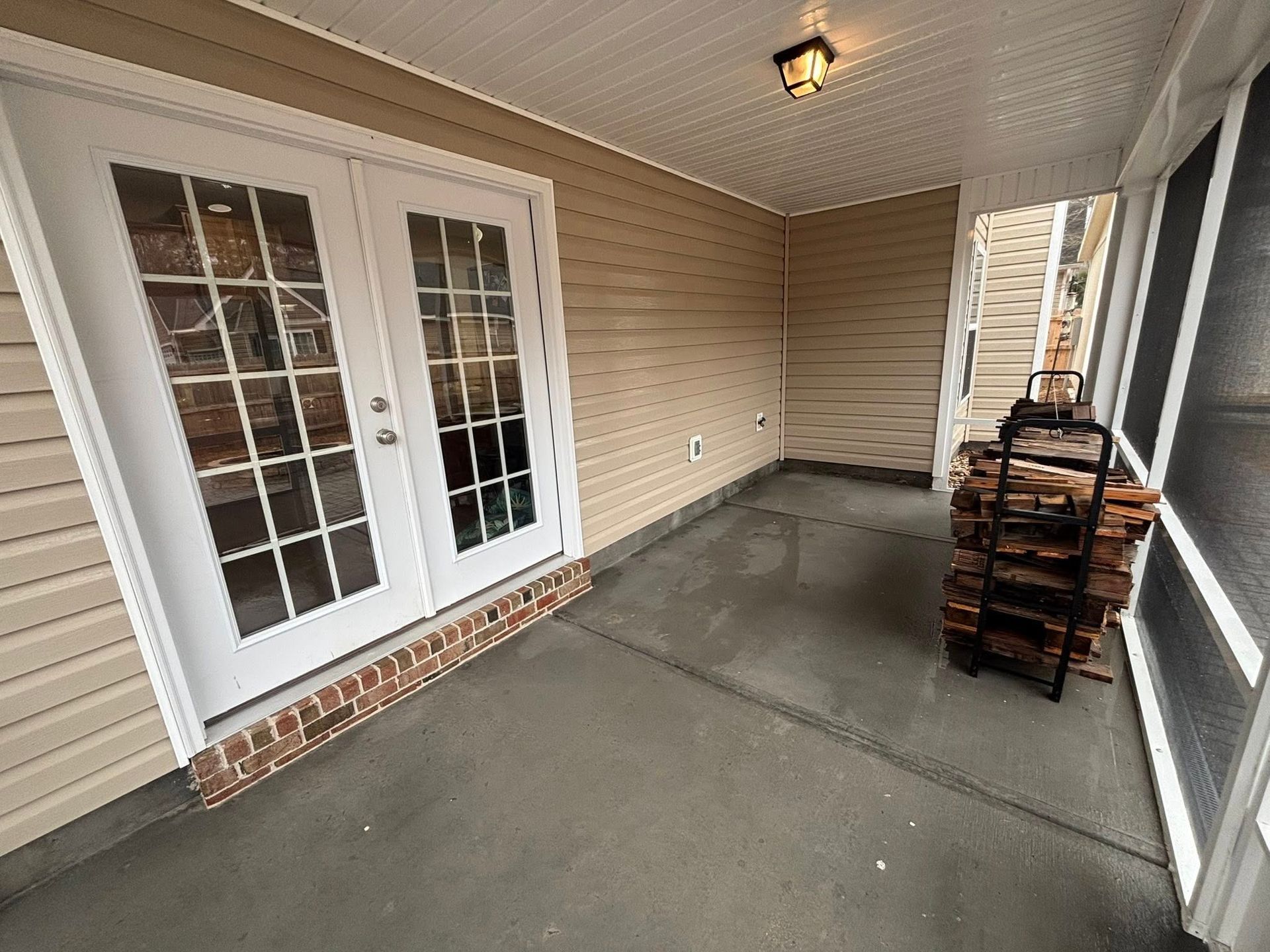 A covered concrete patio with beige siding and a white French door, featuring a black metal rack stacked with firewood.