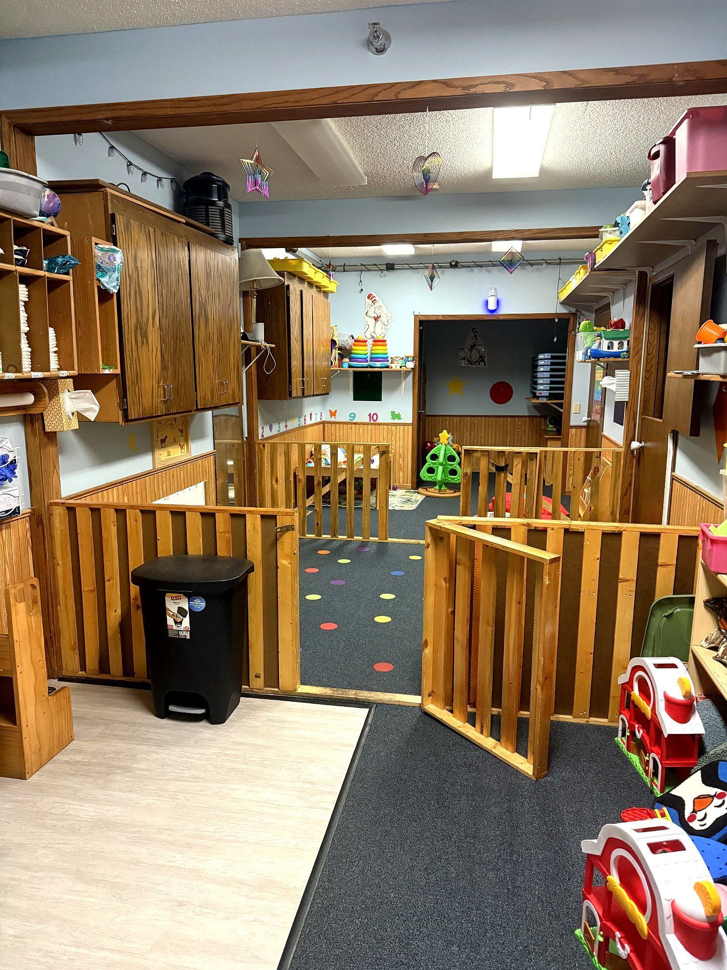 Indoor play area with wooden gates and cubbies. Black trash can, colorful floor and toys visible.
