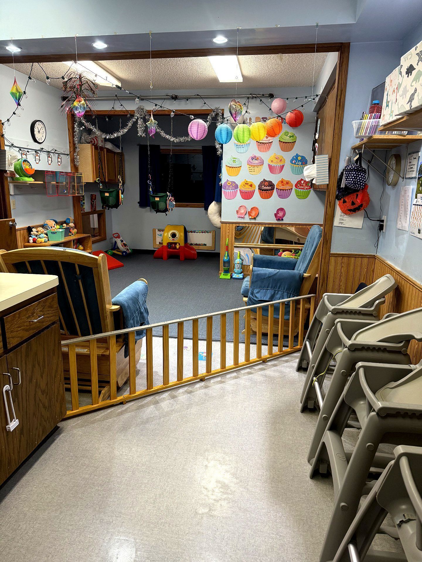 Interior of a colorful daycare. A wooden gate and high chairs lead to a playroom with toys and decorations.