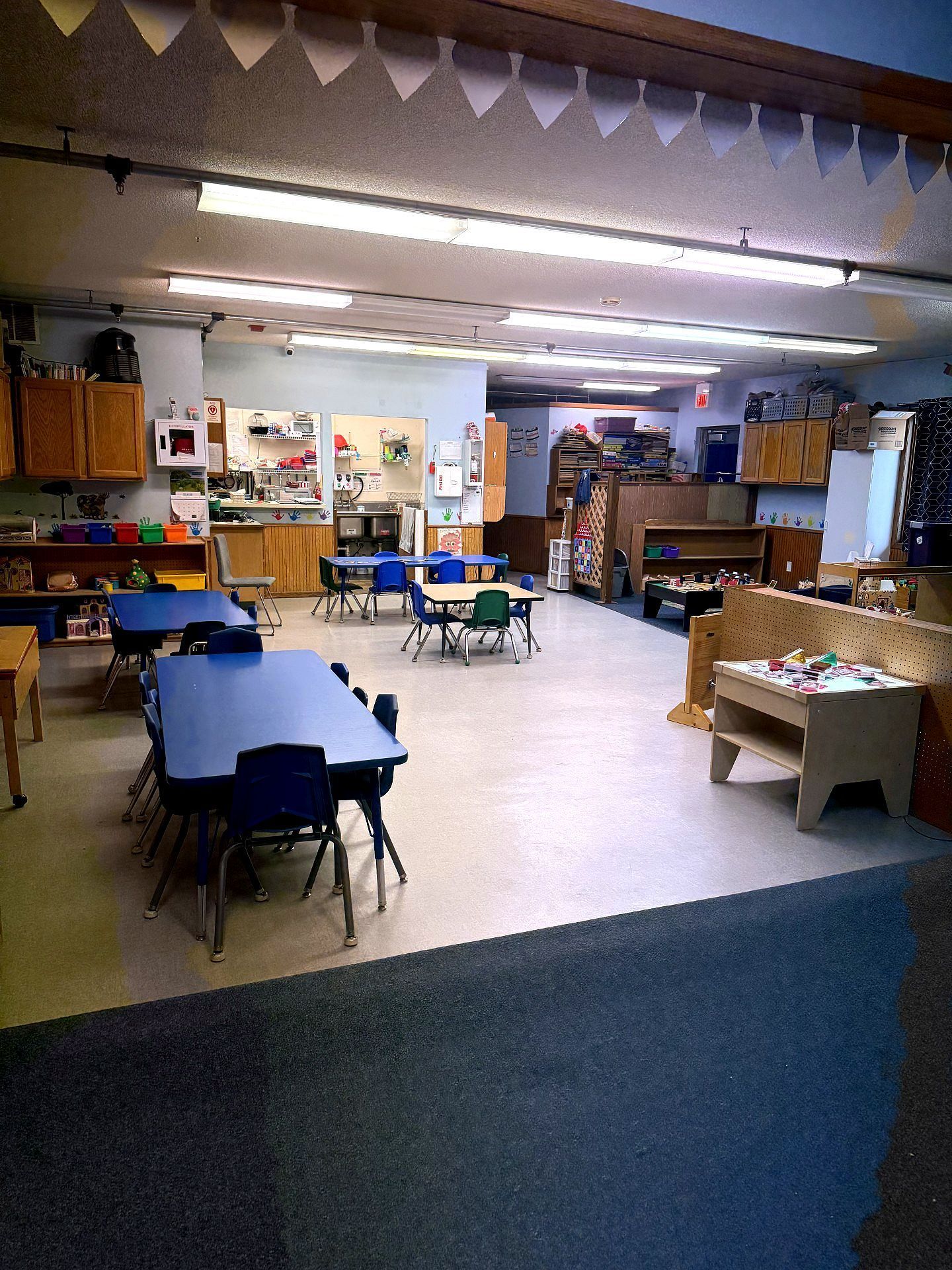 Empty classroom with blue tables and chairs, a sink, and shelving.