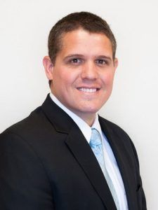 Portrait of a smiling man in a black suit and light blue tie against a white background