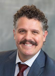 Portrait of a smiling man in a blue suit, white shirt, and maroon tie against a gray background