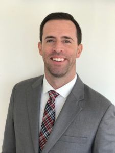Smiling man in a gray suit and red plaid tie against a plain light background