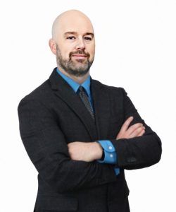 Man in a dark suit with blue shirt and tie, standing with arms crossed against a white background
