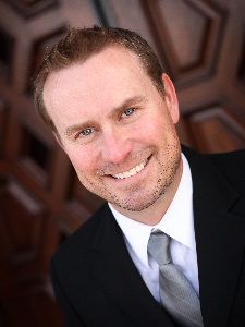 Smiling man in a black suit and silver tie, posed against a dark wooden background.