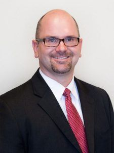 Man in black suit and red tie smiling against a plain light background