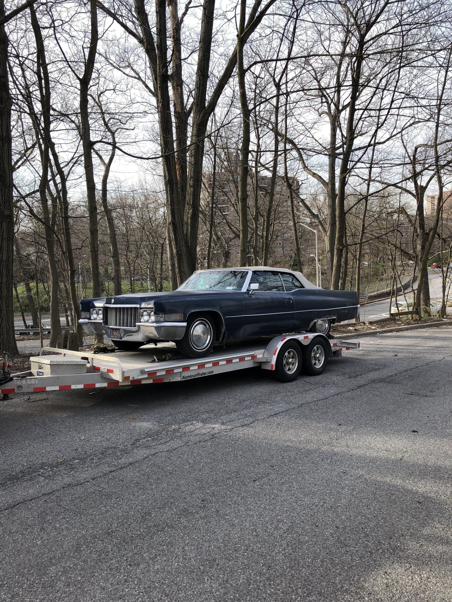Black classic car on a flatbed trailer parked on a gravel road in a wooded area.