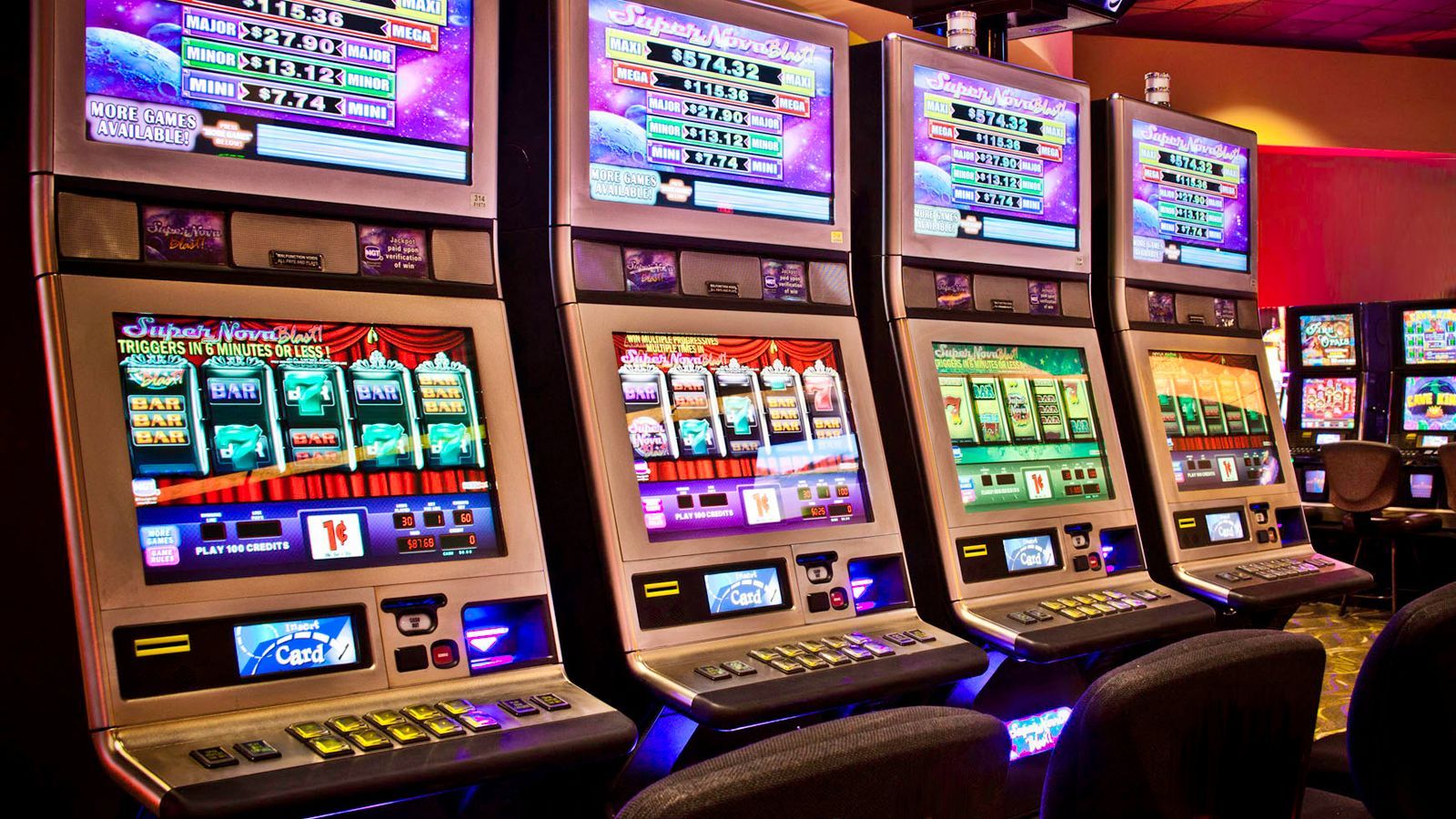 Row of colorful slot machines in a casino lounge with black chairs on a red patterned carpet.