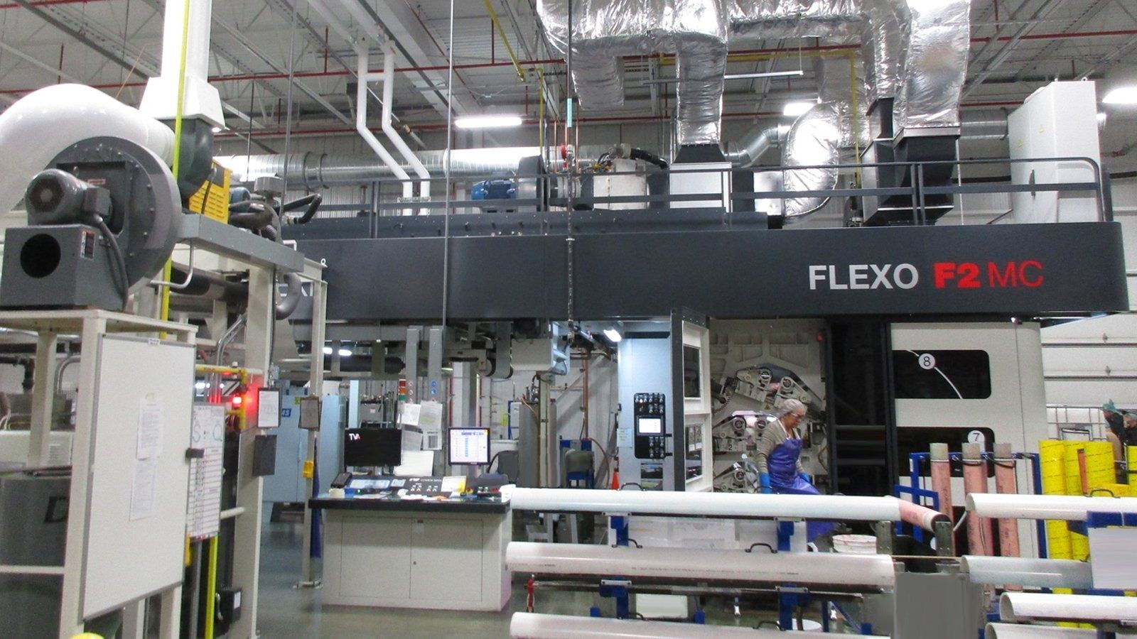 Industrial workshop with a large black FLEXO machine, pipes overhead, and workers nearby