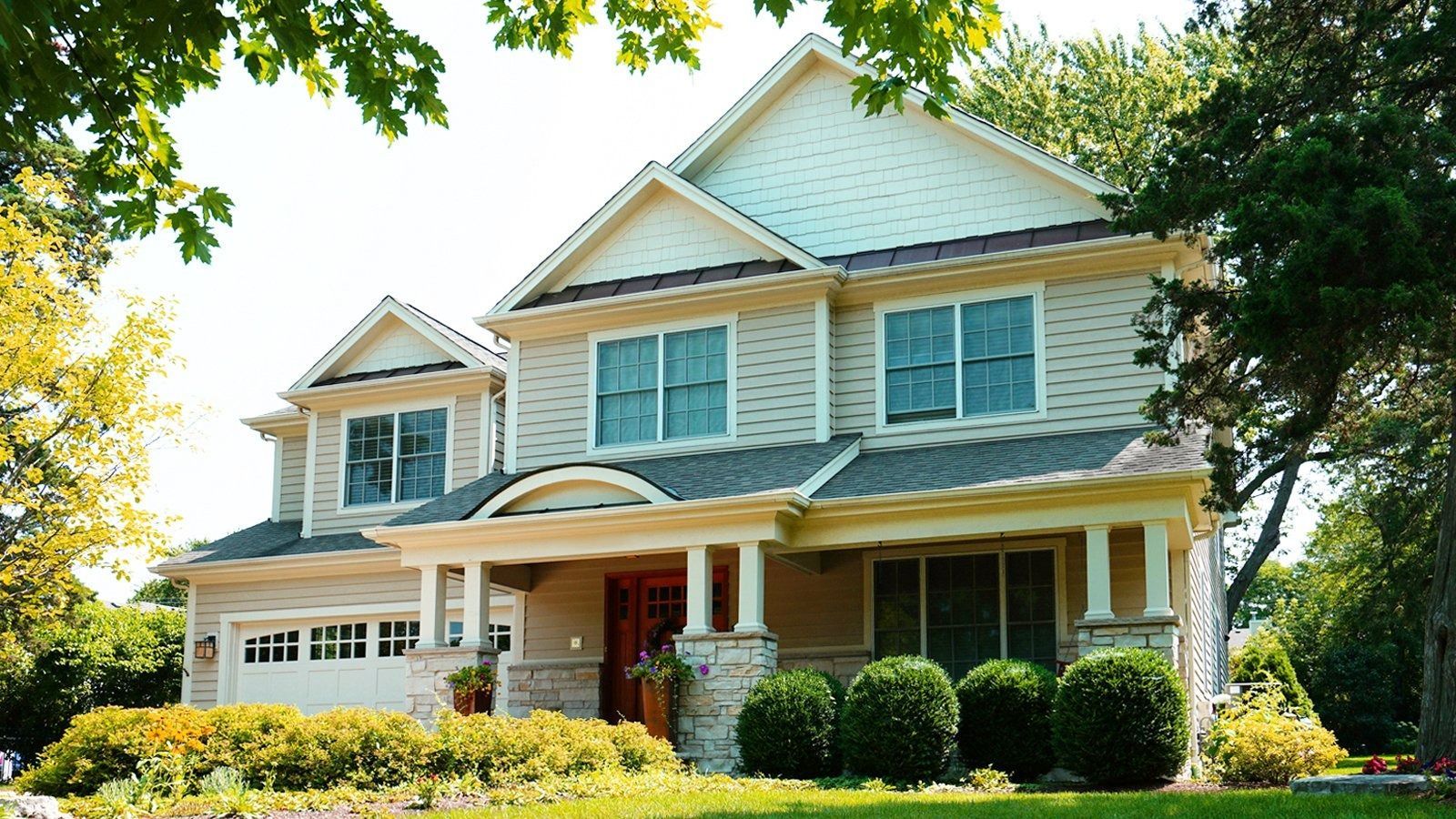 Large two-story beige house with white trim, a front porch, and green trees and shrubs in the yard