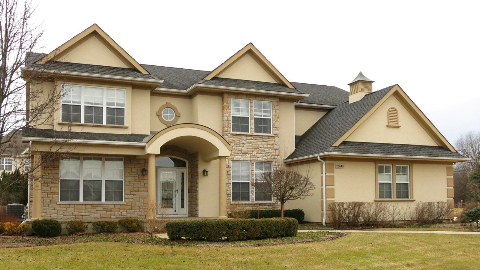 Large two-story beige suburban house with a front porch, stone facade, and manicured lawn