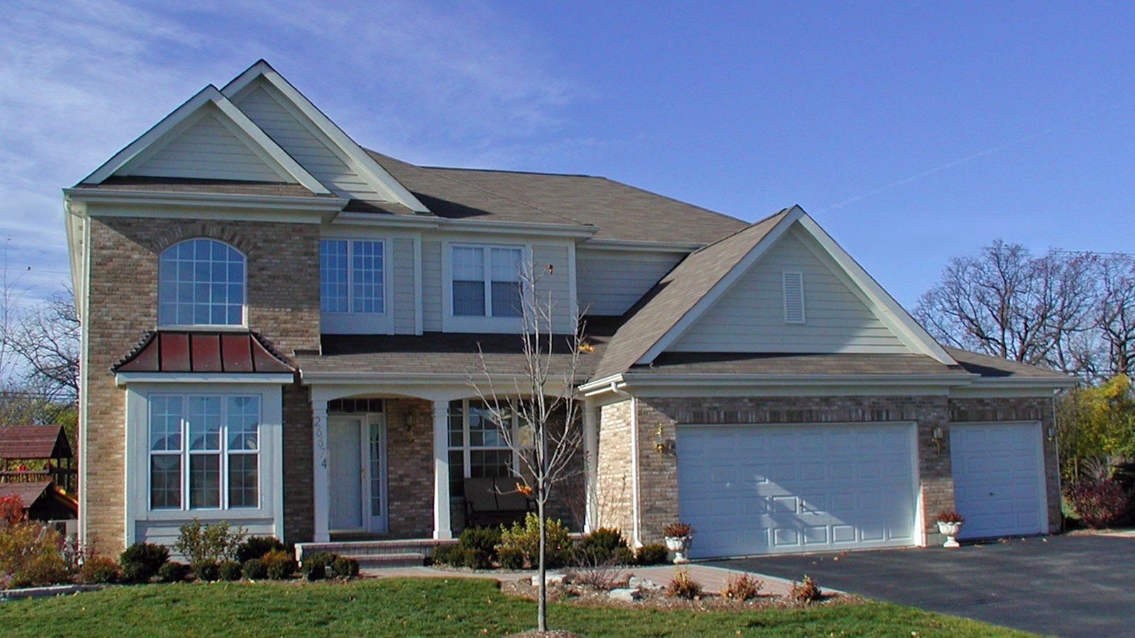 Two-story beige suburban house with a white garage, front porch, and manicured lawn under a blue sky