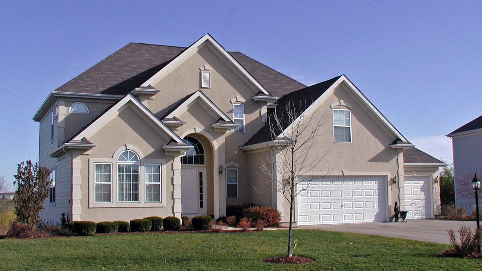 Large beige suburban house with a gabled roof, front lawn, and attached garage on a sunny day