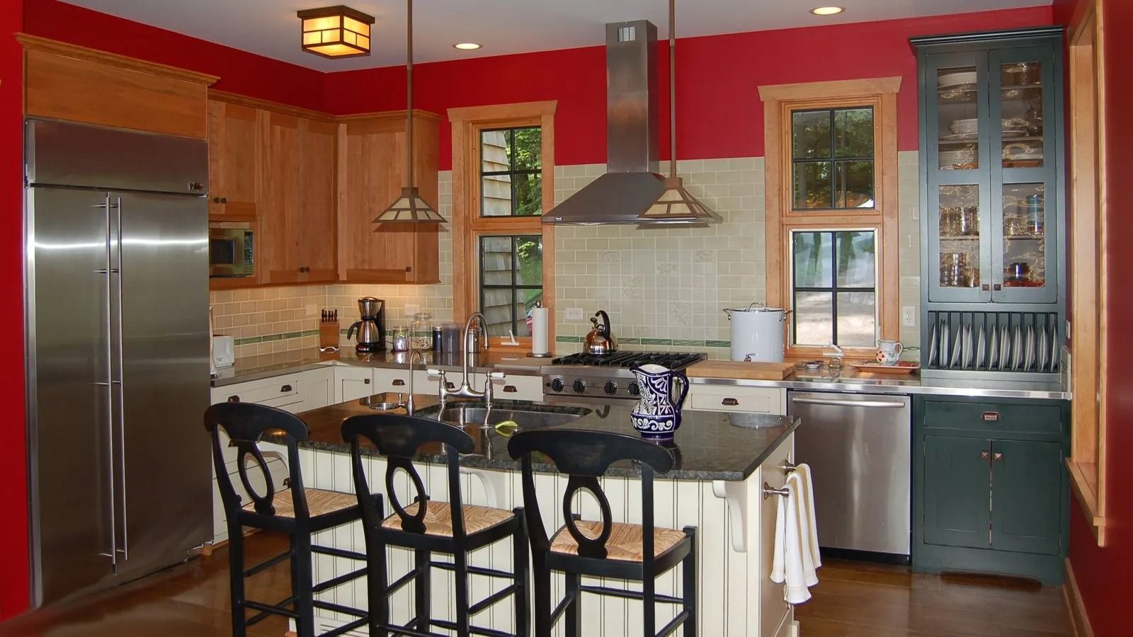 Modern kitchen with red walls, wood cabinets, stainless fridge, island, and black bar stools