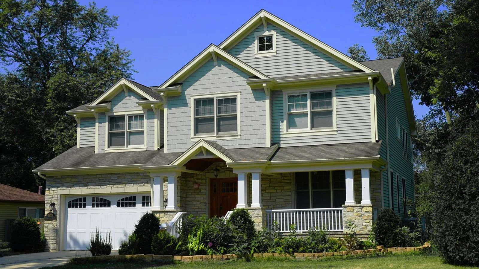 Two-story suburban house with white siding, green trim, and a garage, surrounded by trees and lawn