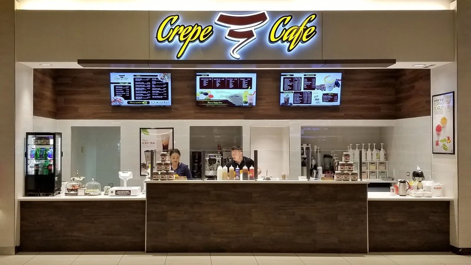 Crêpe Café counter with menu screens, display cases, and a lit sign in a mall food court