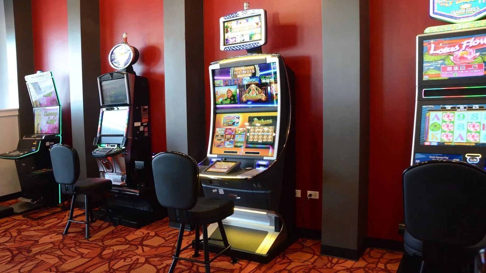 Row of colorful slot machines in a casino lounge with black chairs on a red patterned carpet.