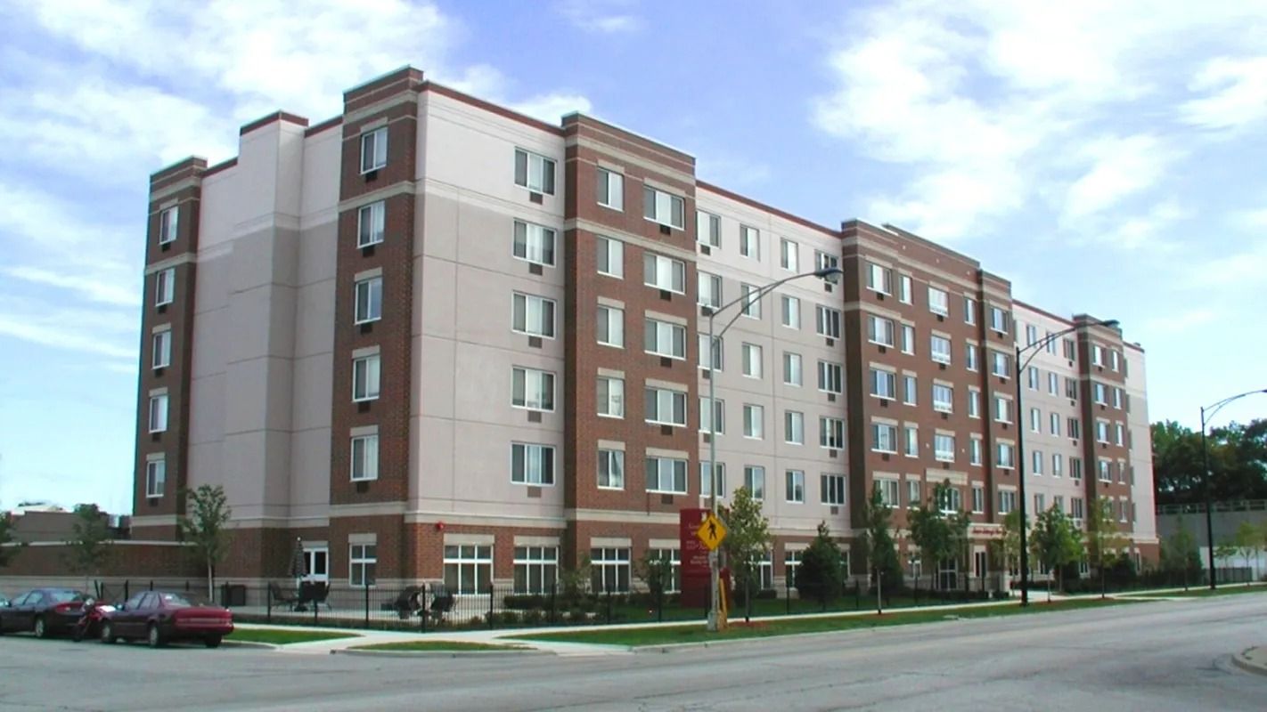 Multi-story brick and white apartment building on a city street, with parked cars and trees nearby