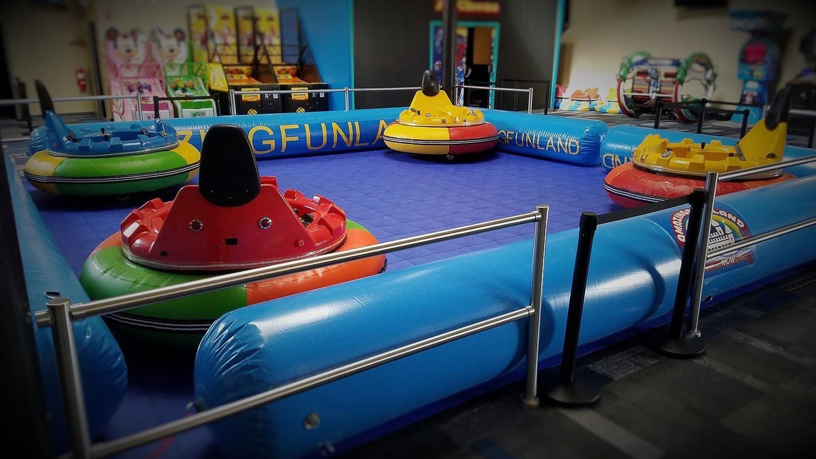 Indoor bumper car arena with colorful cars on a blue ride platform, viewed through metal railings.