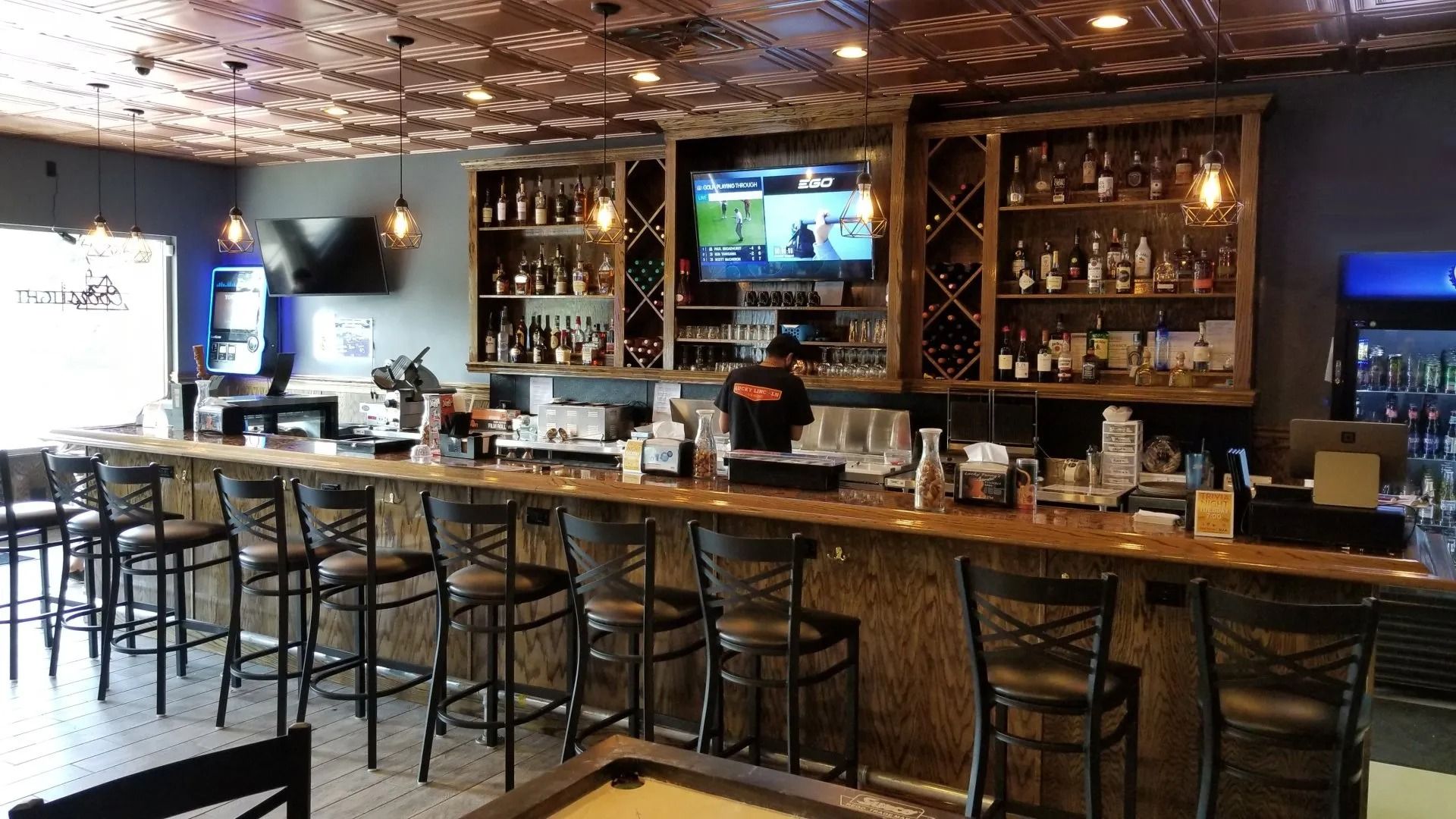 Cozy bar interior with high stools, wooden shelves, hanging lights, and a TV above the counter.