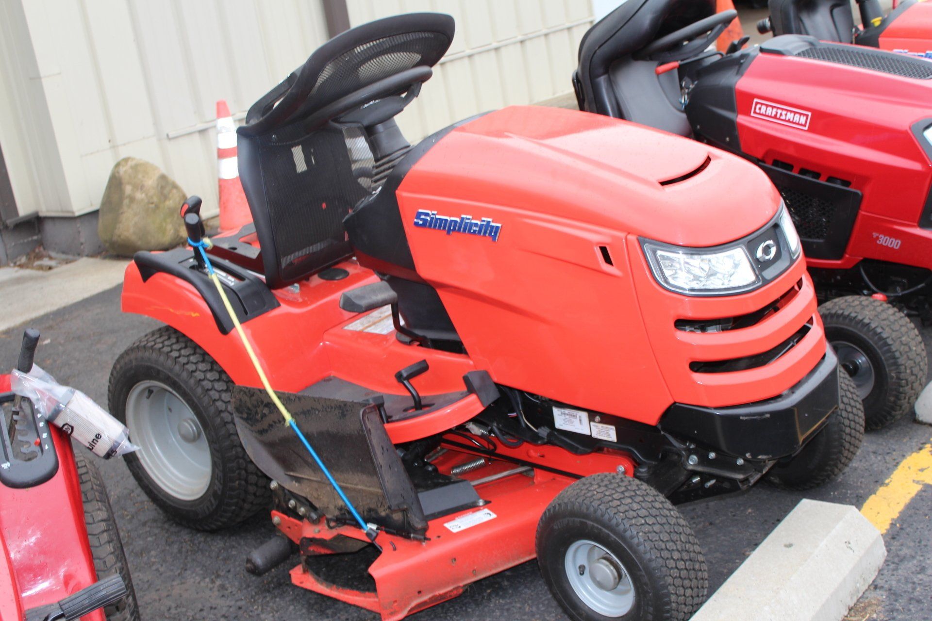 Red Simplicity riding lawnmower on pavement, outdoors. Another red mower is visible in the background.