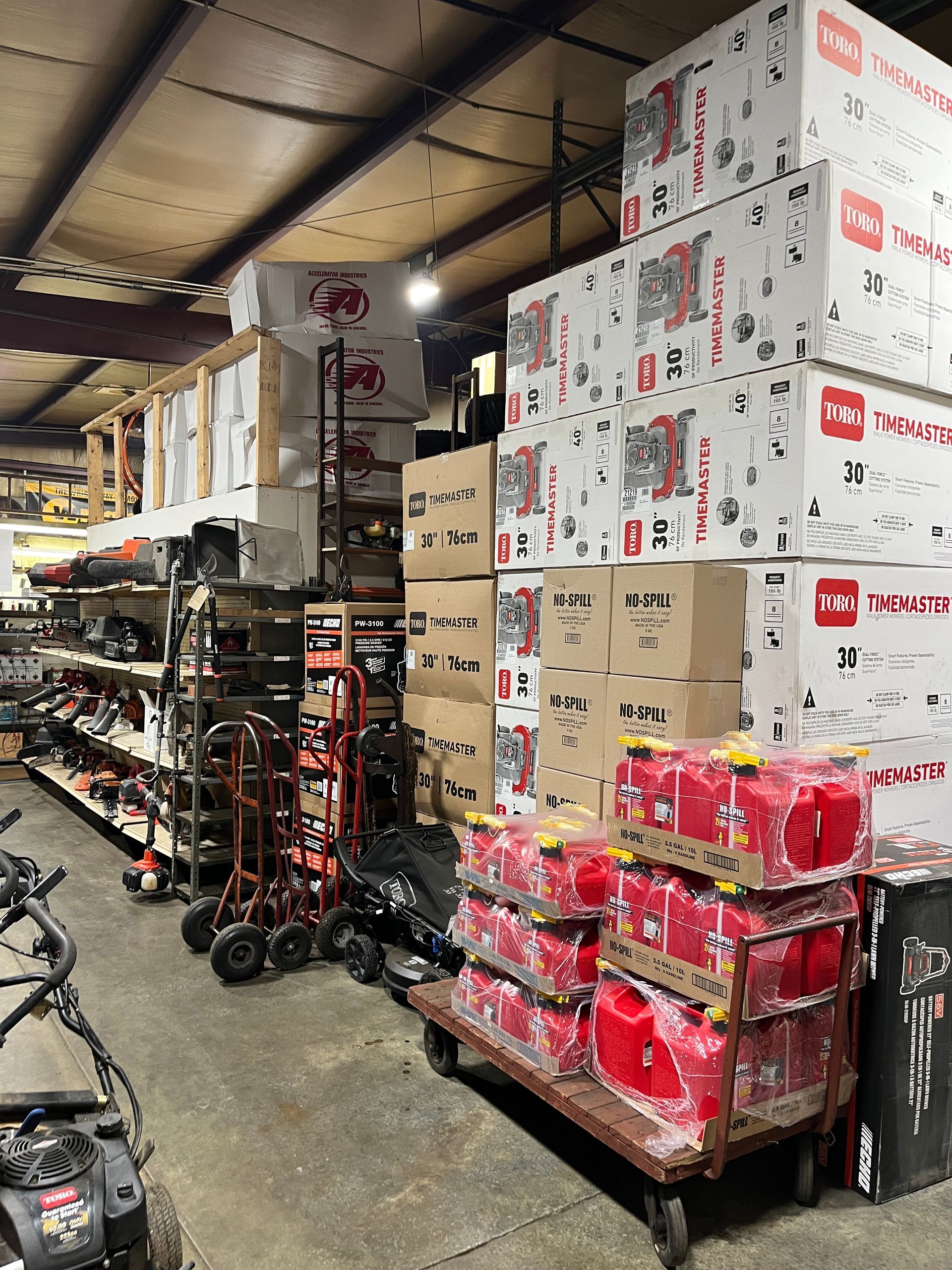 A warehouse aisle filled with stacked boxes of outdoor equipment, shelving units, and red fuel cans on a rolling cart.