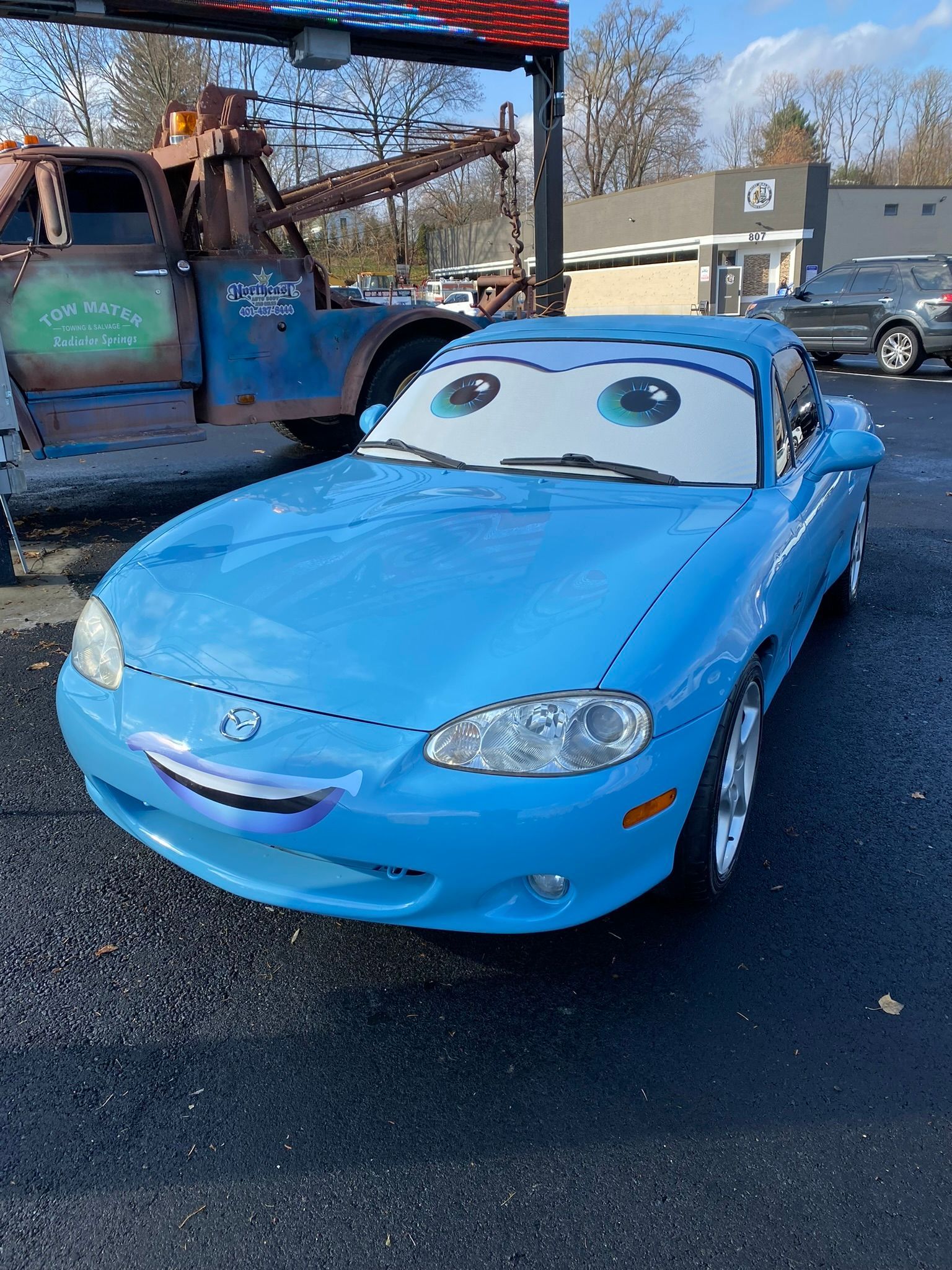 A blue car with a tow truck in the background is parked in a parking lot.