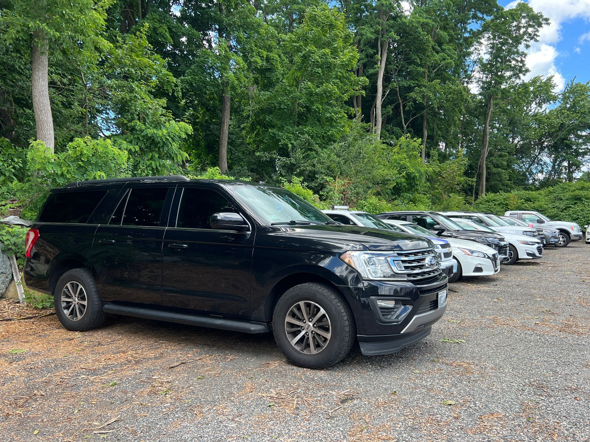 A row of black cars are parked in a gravel lot.