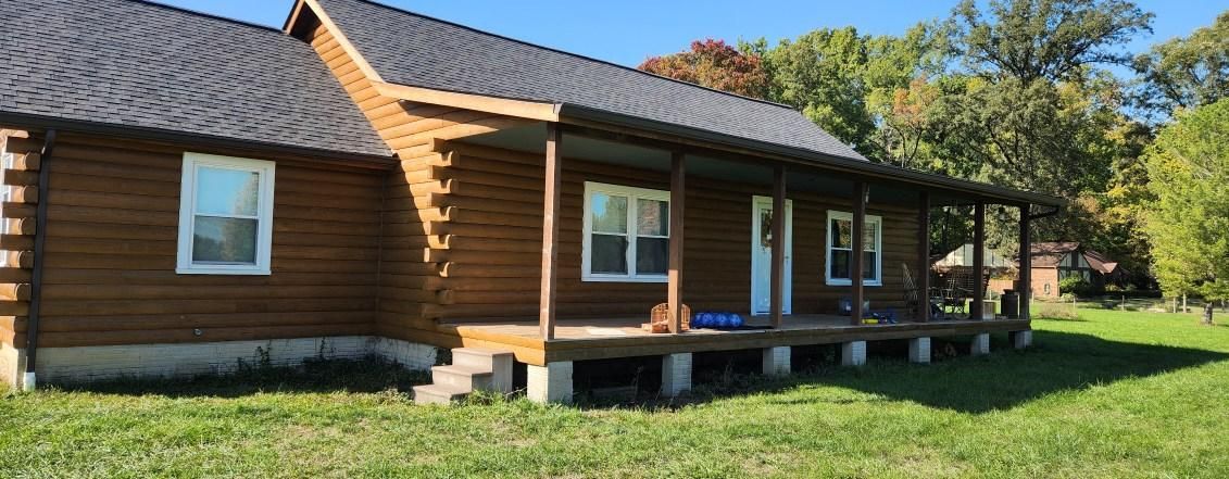 Log cabin with wooden porch and windows, set in a sunny grassy yard with trees nearby