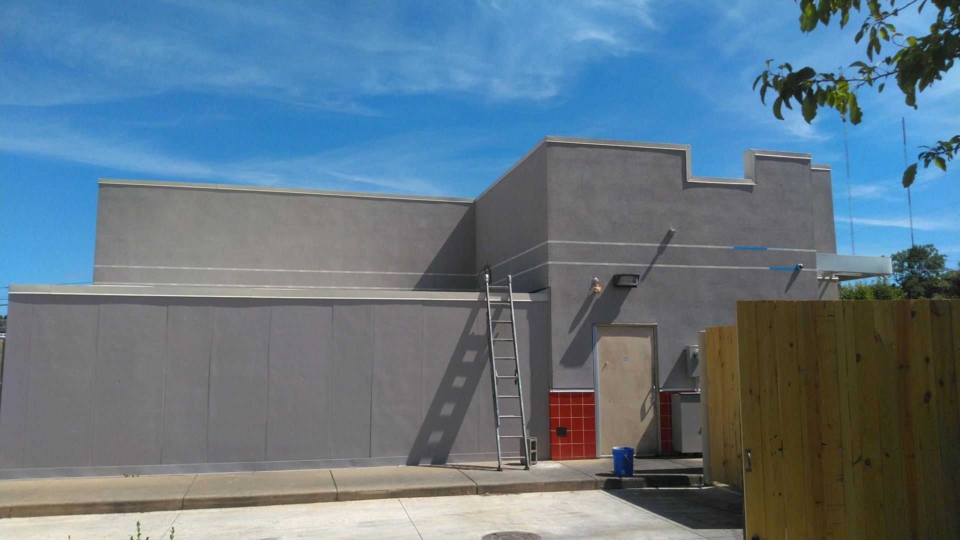 Gray stucco building with a red door and ladders leaning against the wall under a blue sky