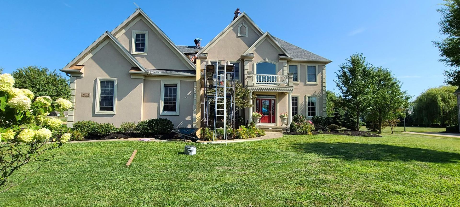 Large beige suburban house with a manicured lawn, flower beds, and a clear blue sky