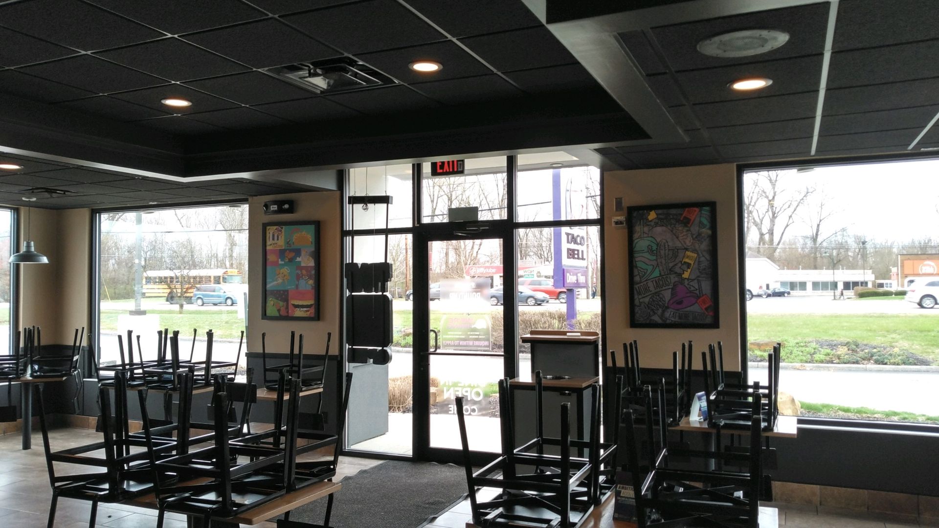 Empty restaurant interior with stacked chairs, tables, and large windows facing a parking lot.