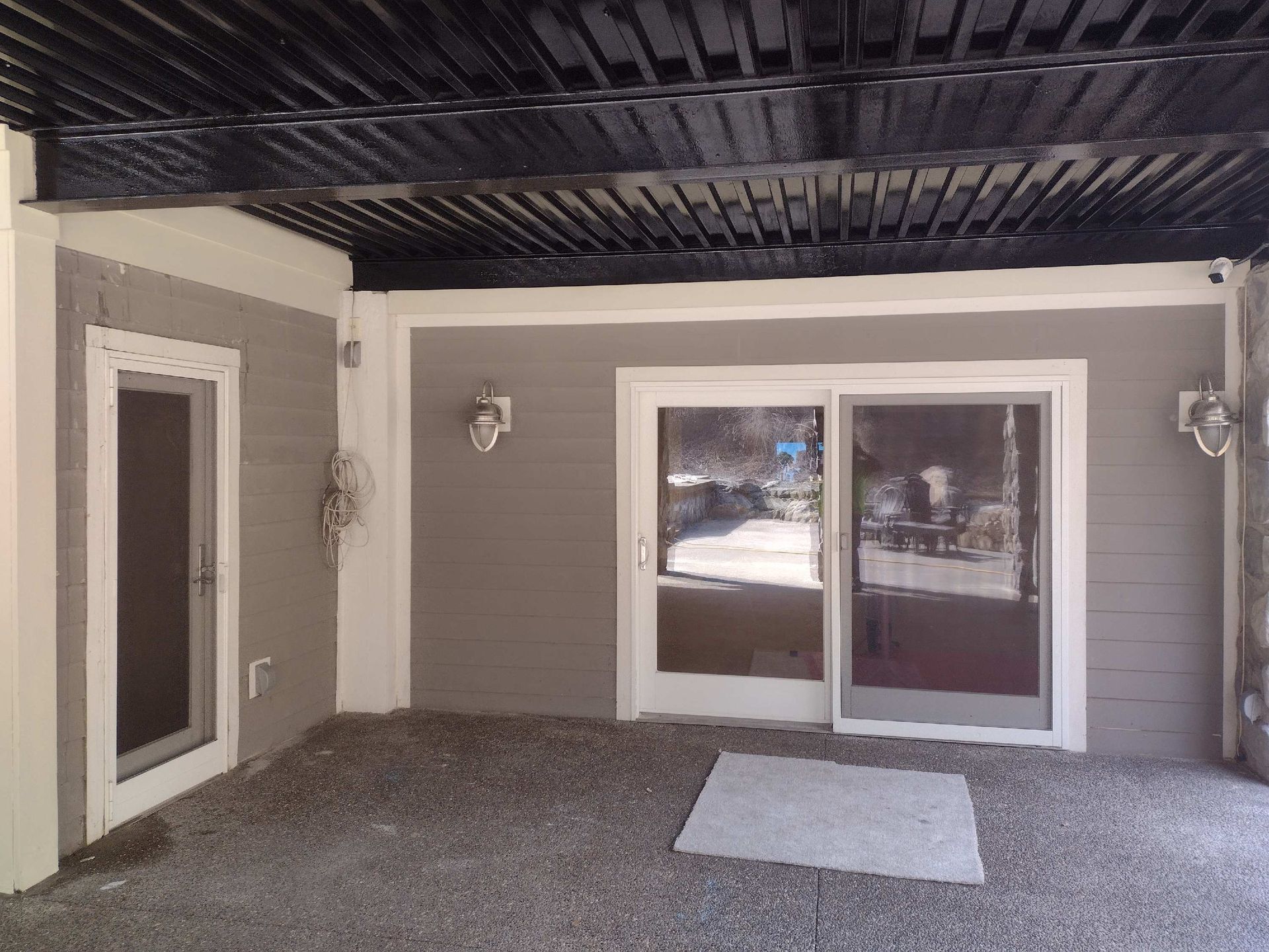 Covered patio with gray siding, dark ceiling, sliding glass doors, and gravel floor.