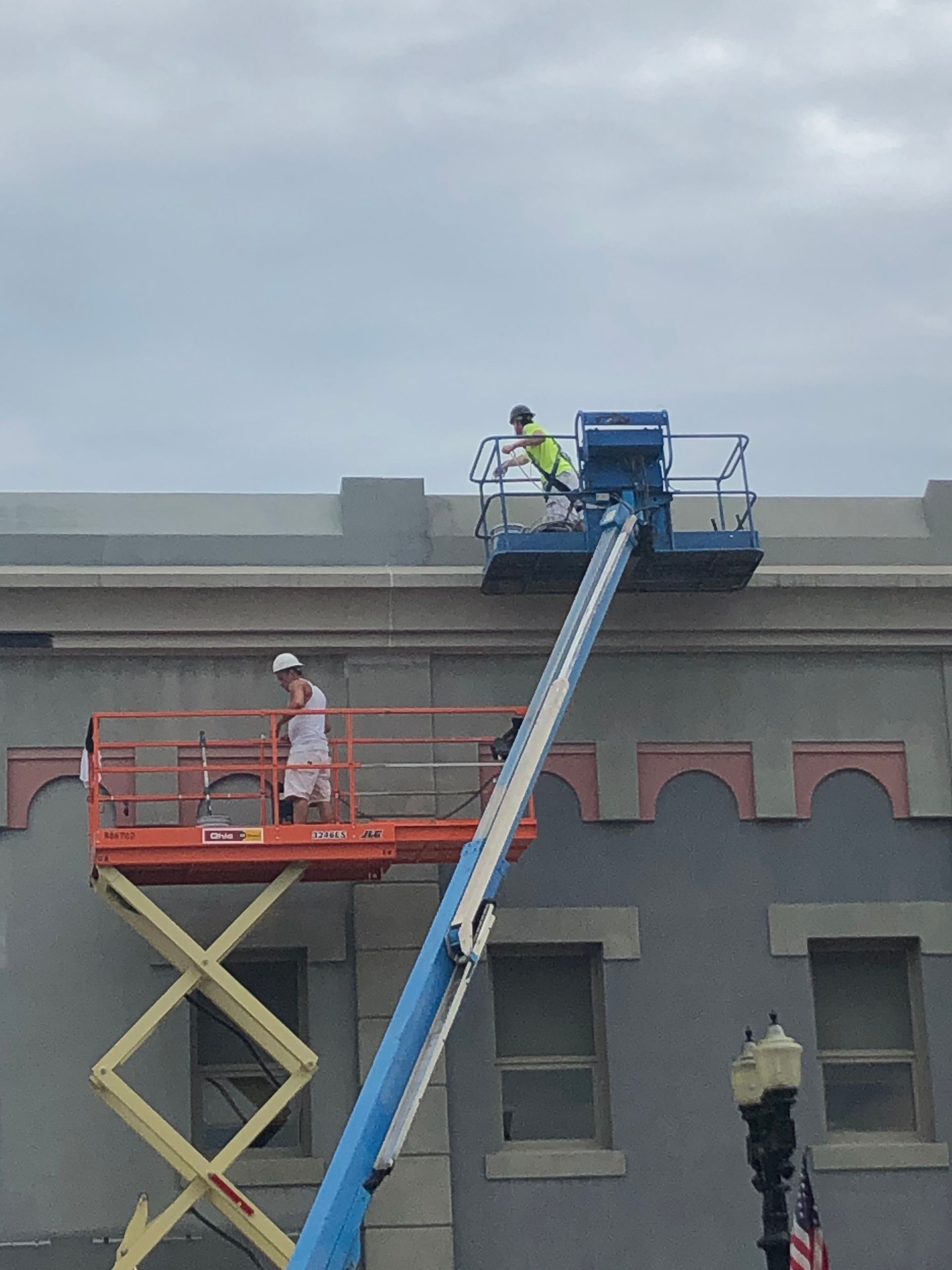 Two workers on lifts painting a gray building roofline under an overcast sky.