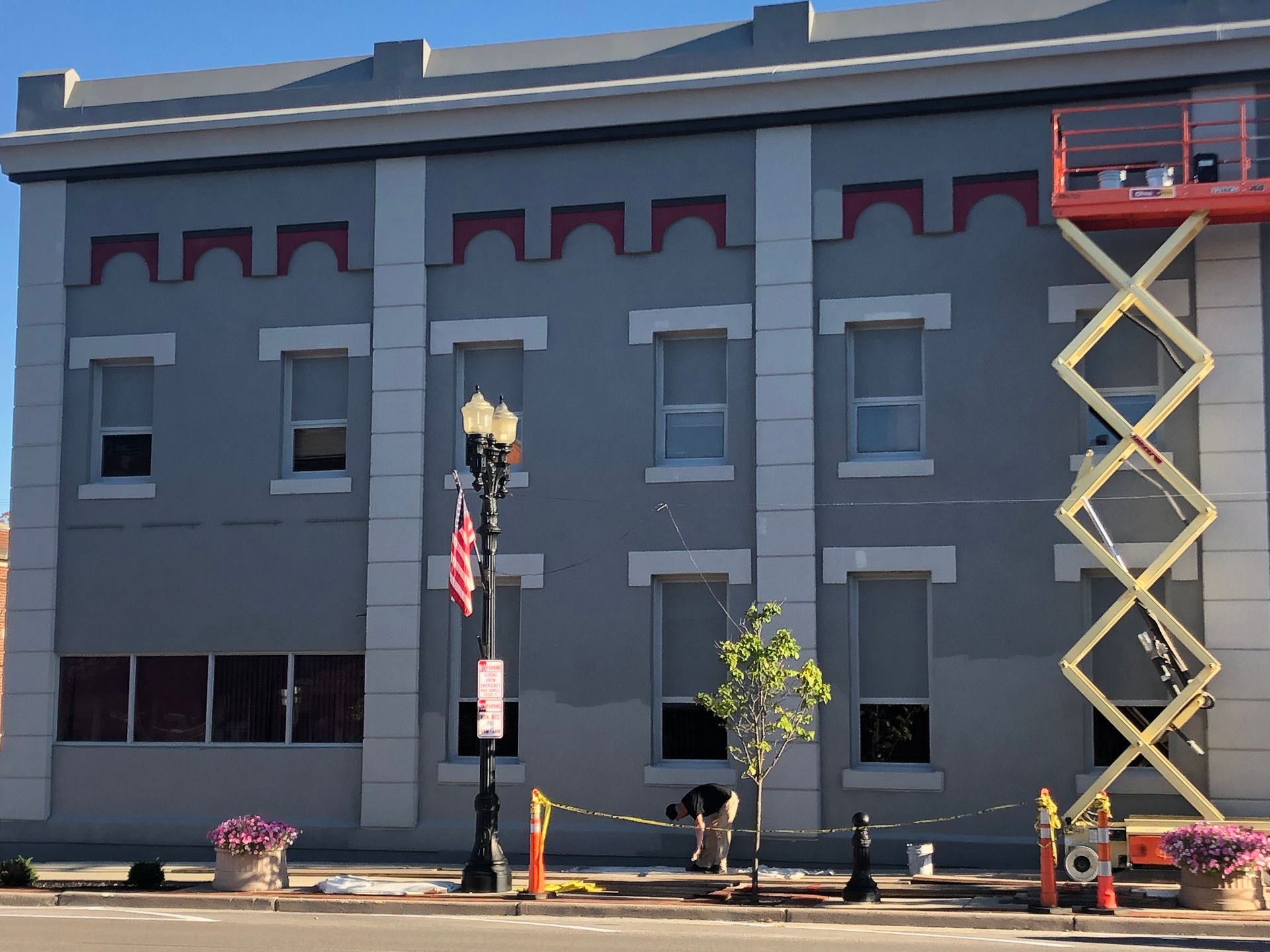 Gray building facade with small windows, an American flag, and a yellow scissor lift at the right side