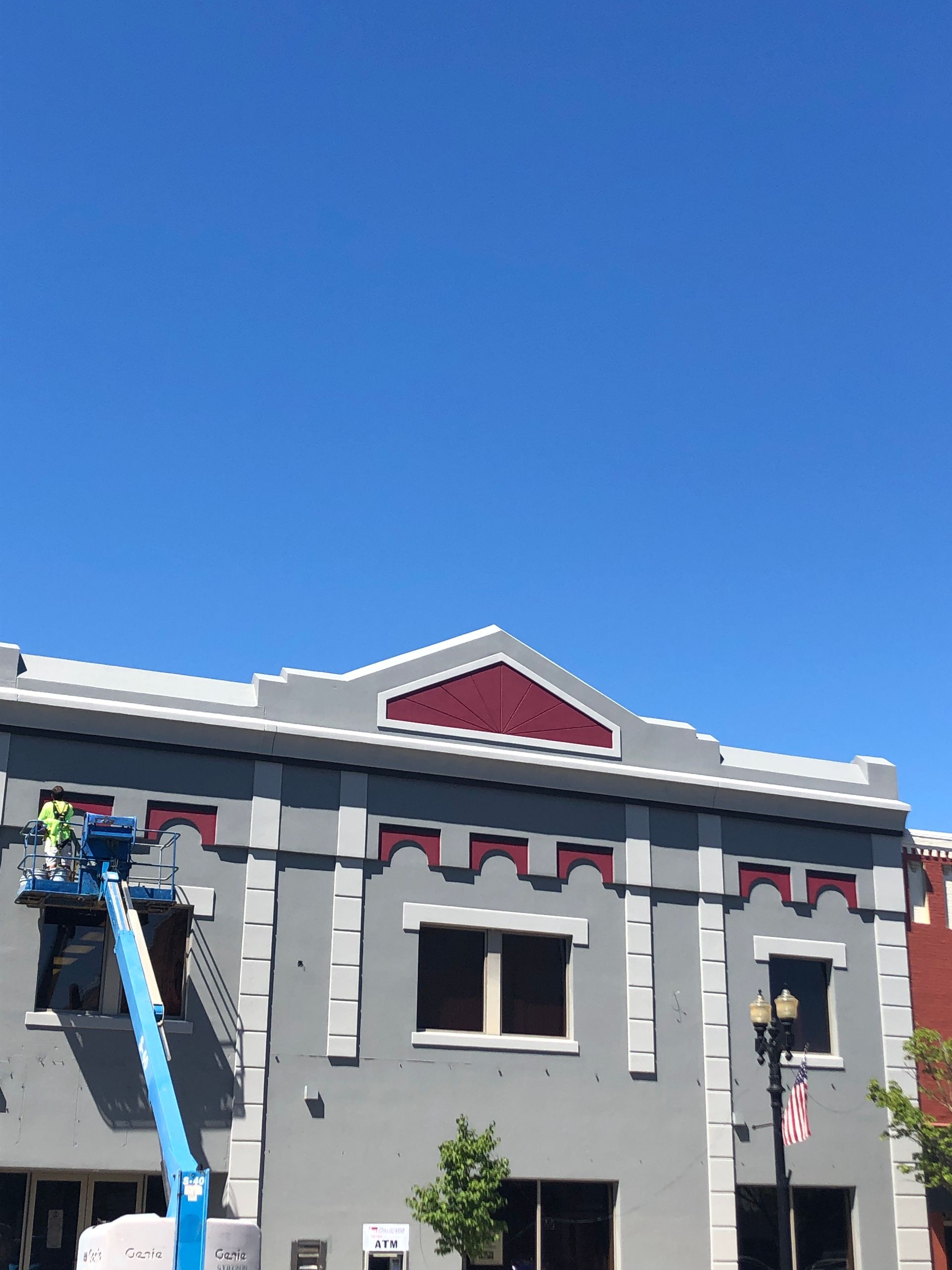 Gray building with red trim under a clear blue sky, with a worker on a blue lift at the left side.