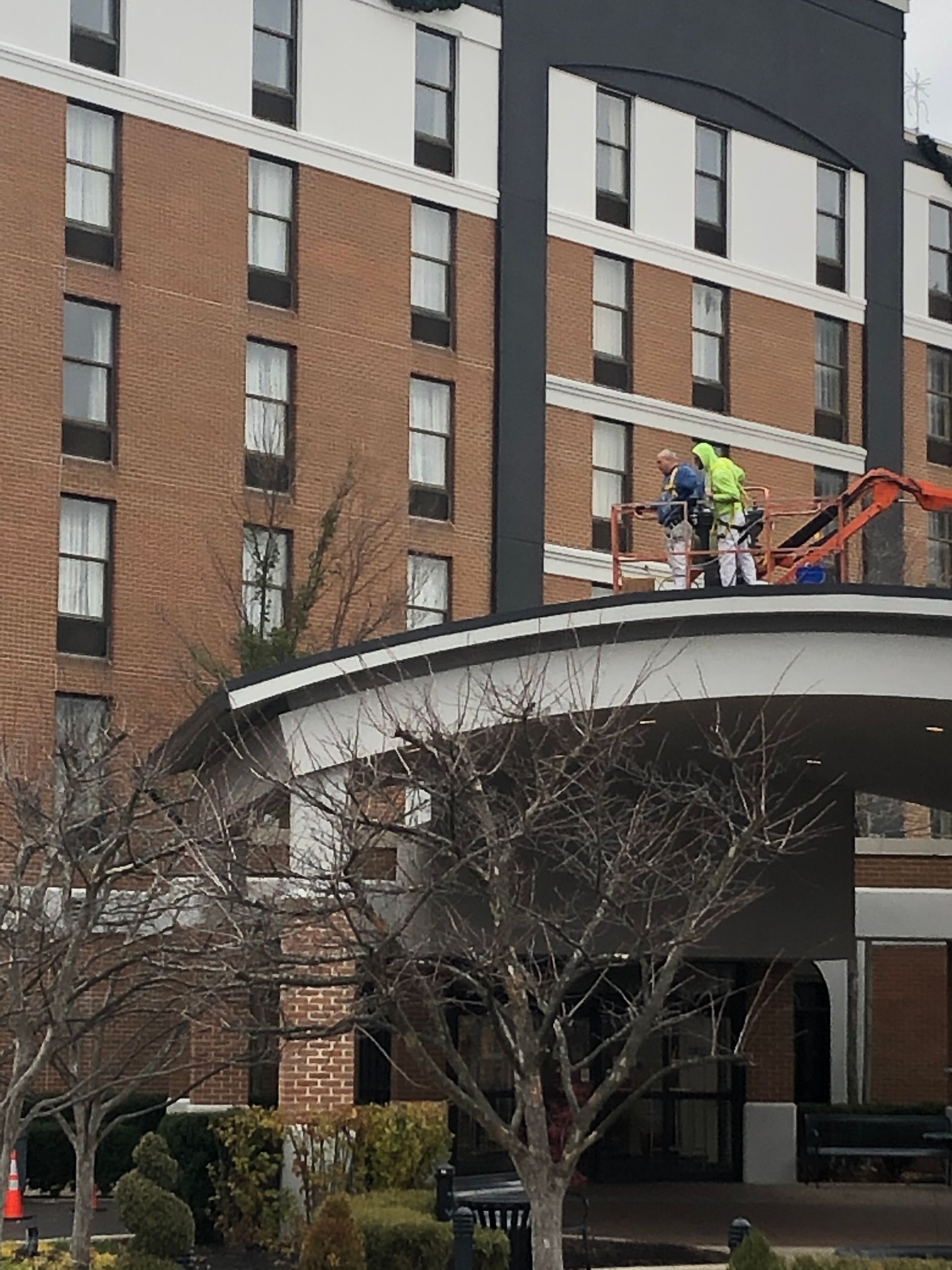 Workers on a rooftop above a building entrance, with a cherry picker and brick apartment facade.