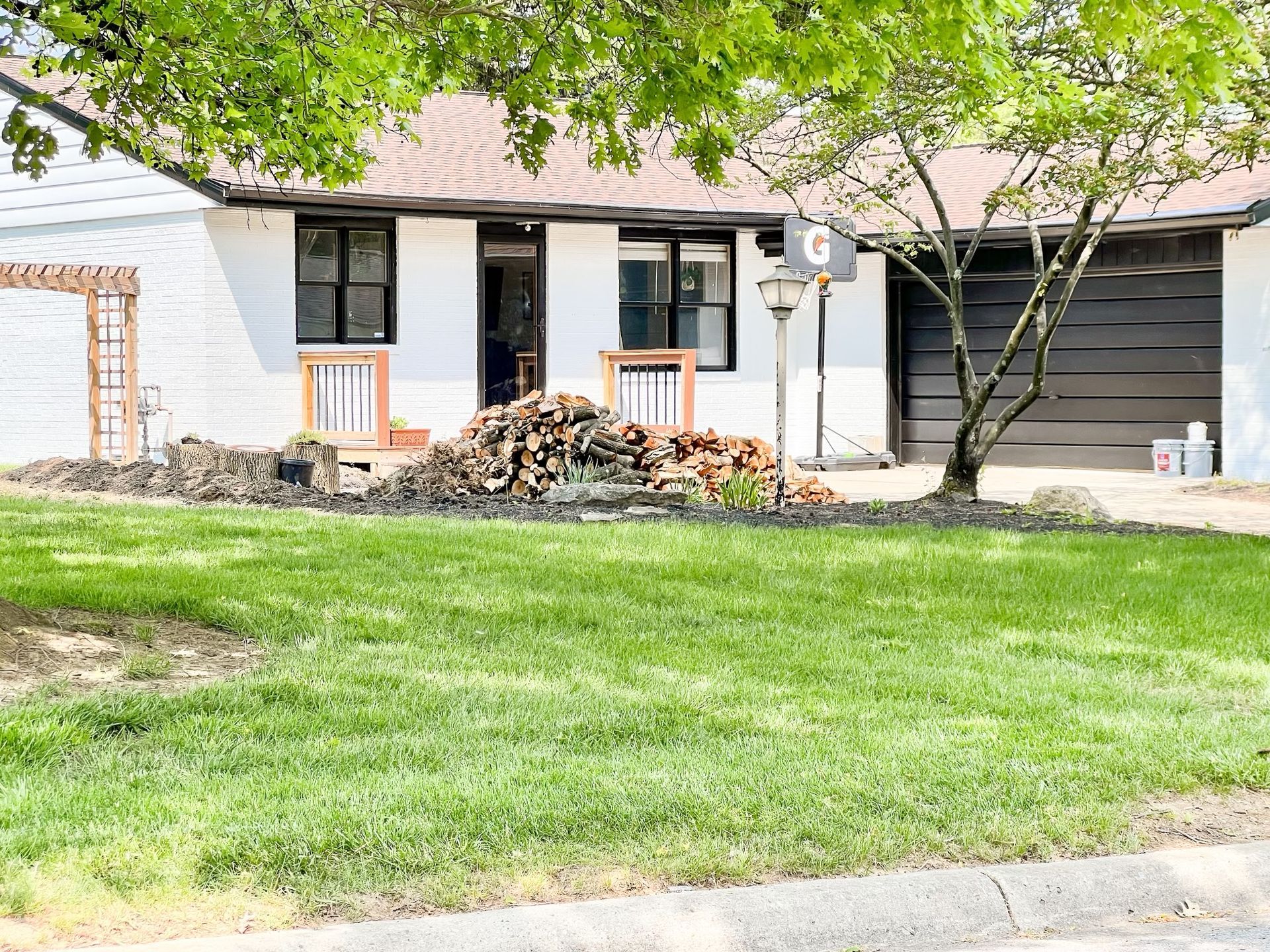 Suburban house with a green lawn, trees, and a pile of firewood by the front porch.